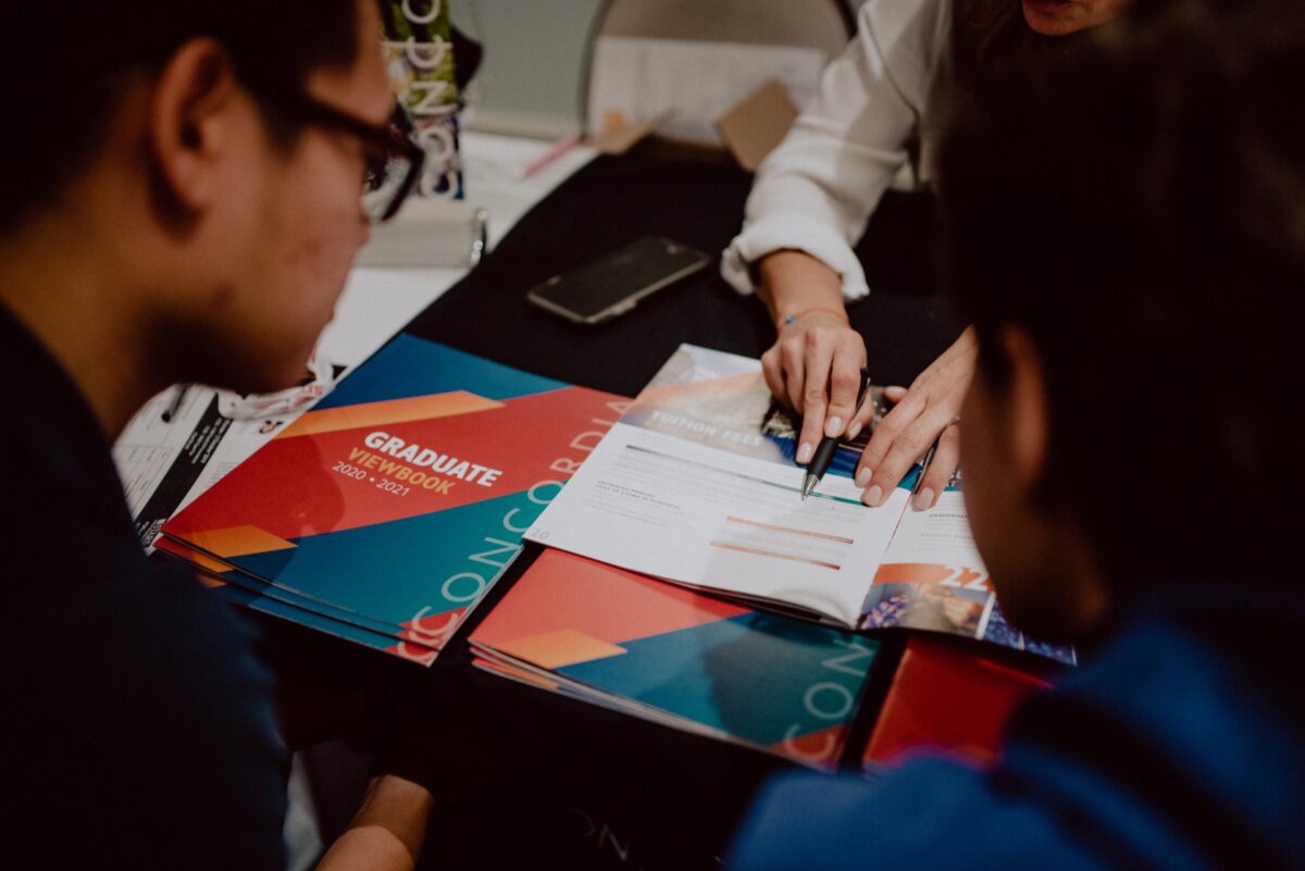 Two people are sitting at a table, looking through a brochure titled "Graduate Viewbook 2020-2021," while a third person points to the pages. There are various handouts on the table, and a smartphone is placed near the brochures. This scene was expertly captured by an event photographer Warsaw to be included in his event photo portfolio.  