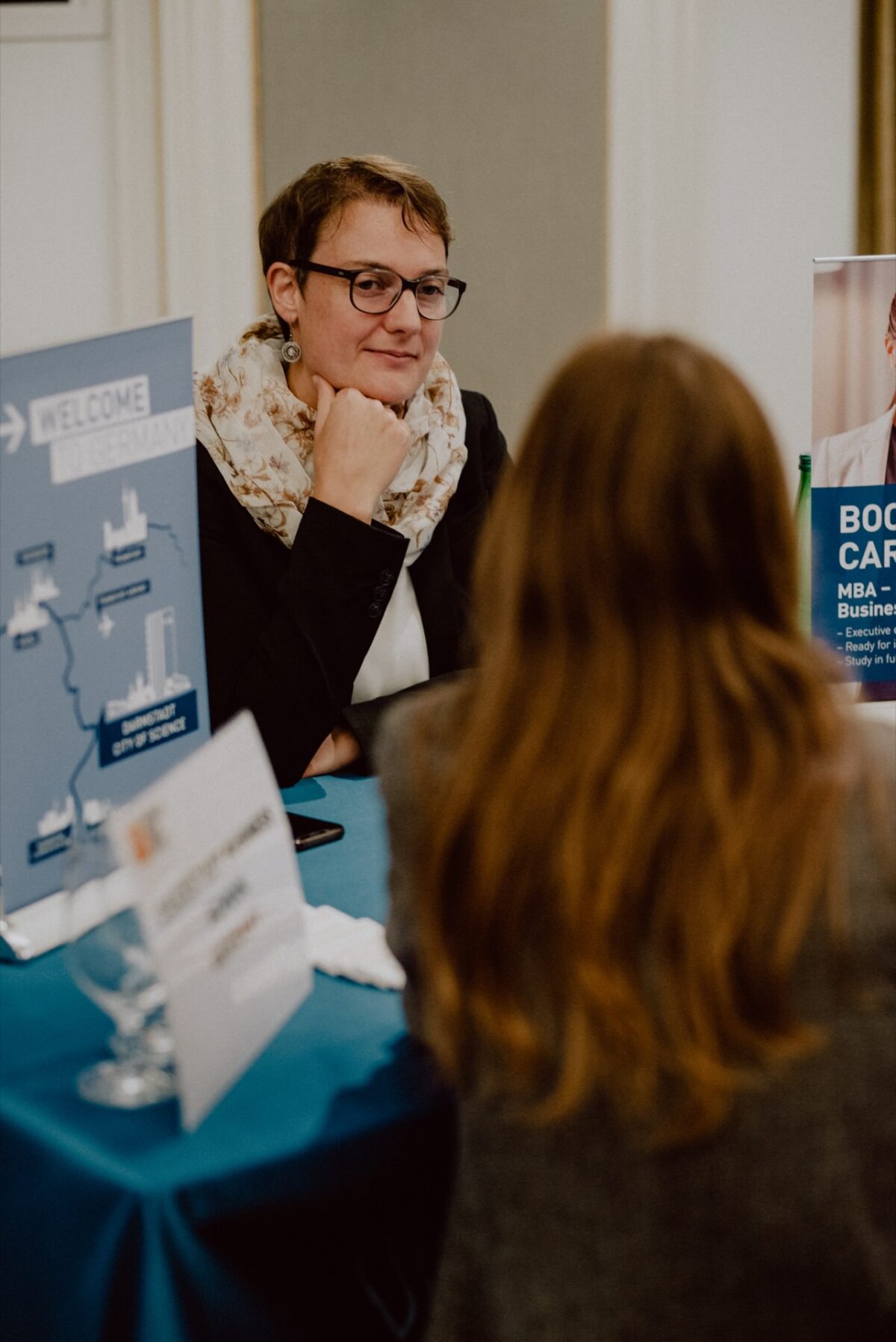 A person with short hair, glasses and a scarf sits at a table and looks busy talking to another person, who is standing with his back to the camera. On the table lies a "Welcome" sign and a business brochure suggesting a professional or networking event. This scene could easily be part of an event photo essay by an event photographer Warsaw.  