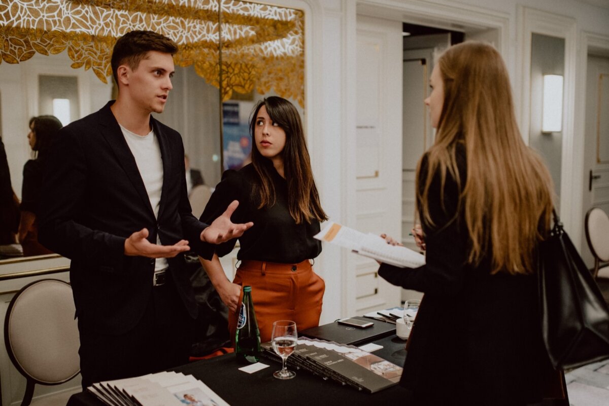 Three people are having a conversation at a professional event. A man and a woman are standing behind a table with brochures and drinks and talking to another woman holding a brochure. The setting, ideal for event photography, is reminiscent of a conference or networking event.  