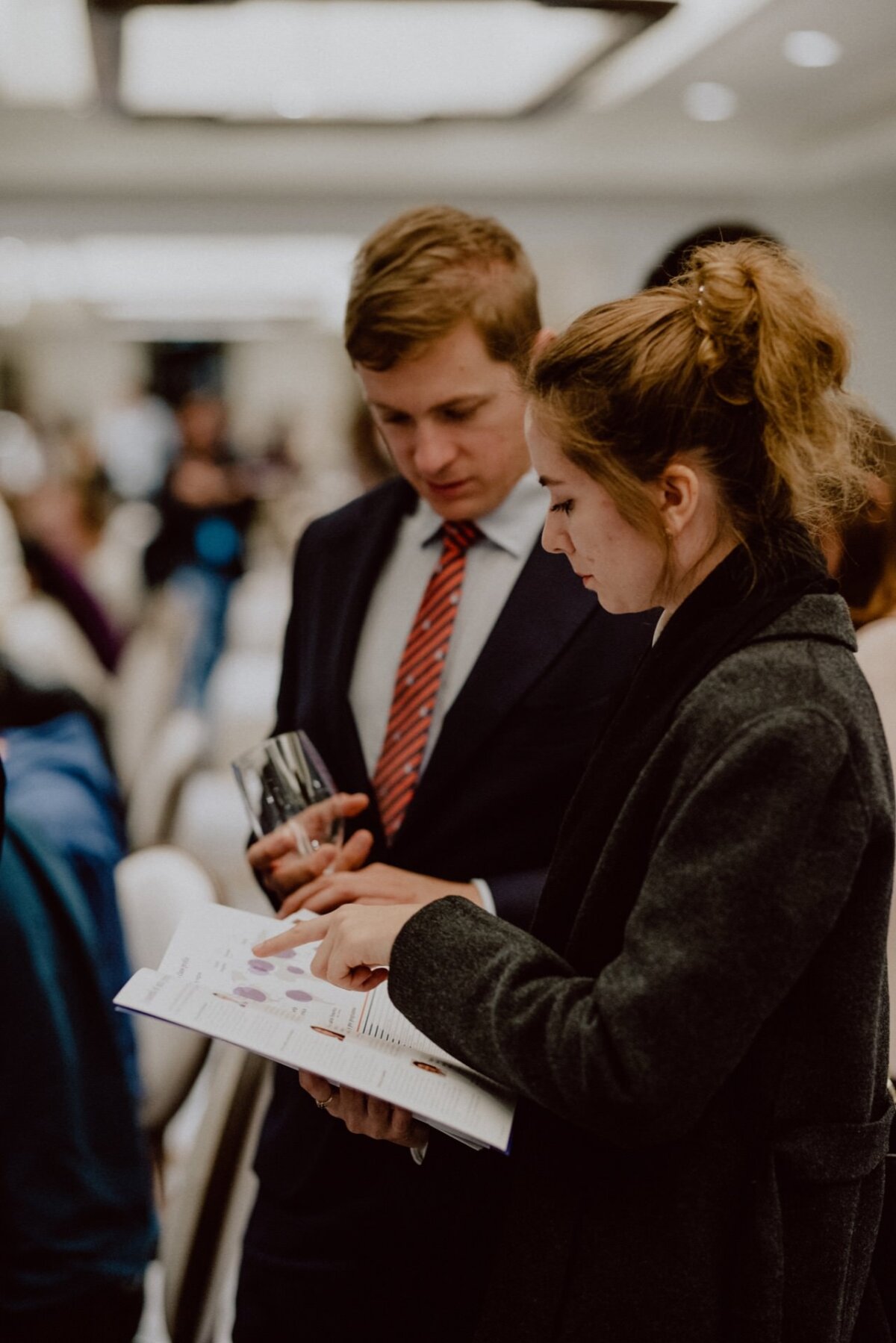 Two people, a man in a red-striped suit and tie and a woman in a dark coat, are standing close together in a softly lit room with many blurred figures in the background. They are looking at a document held by the woman, looking engrossed in conversation - a perfect scene for event photography. 