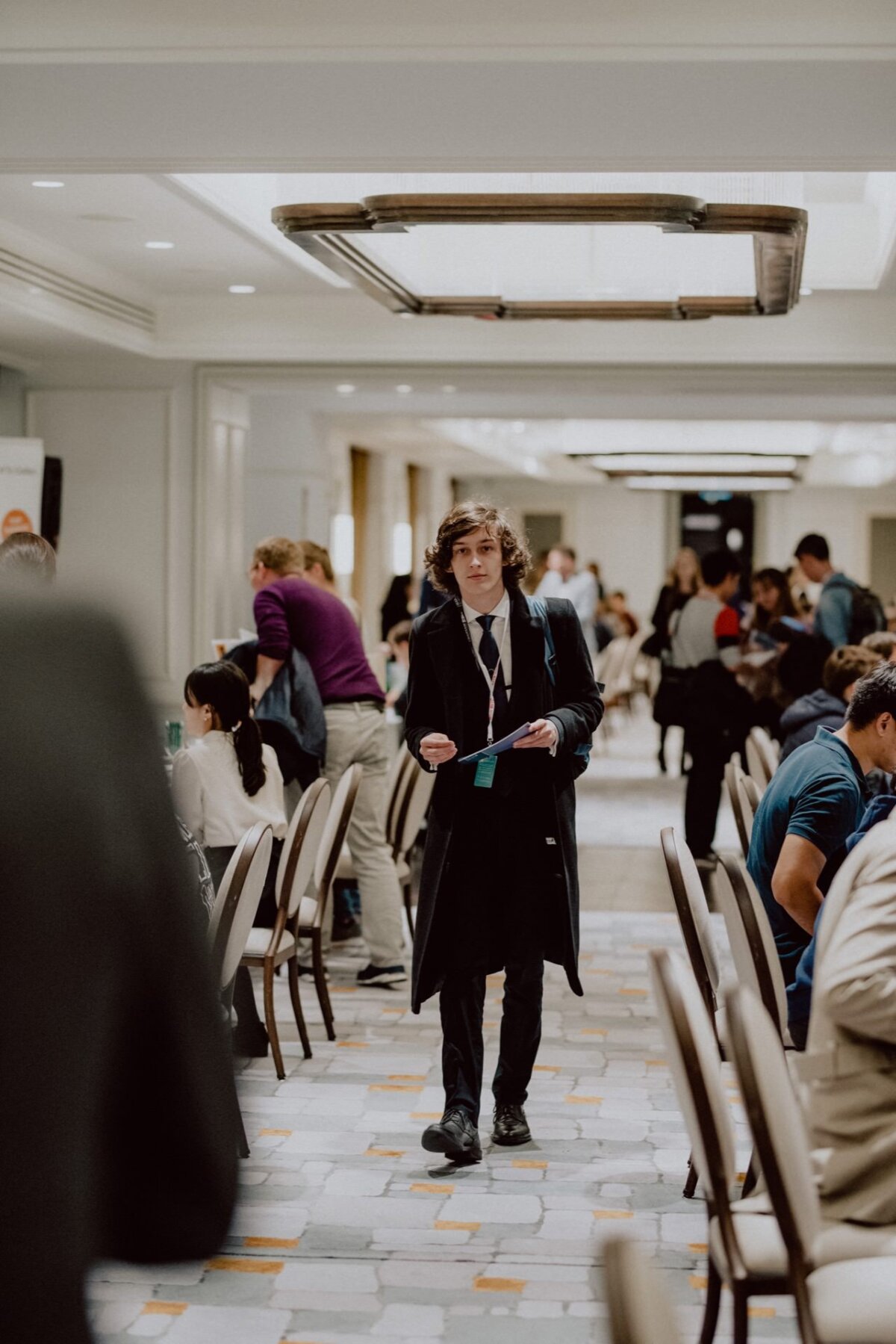 A young man with curly hair, dressed in a dark suit and tie, walks through a room filled with people sitting at tables. Around his neck he holds a paper and a lanyard. The room has a modern interior with overhead lighting and a tense, professional atmosphere - perfect for event photography.  