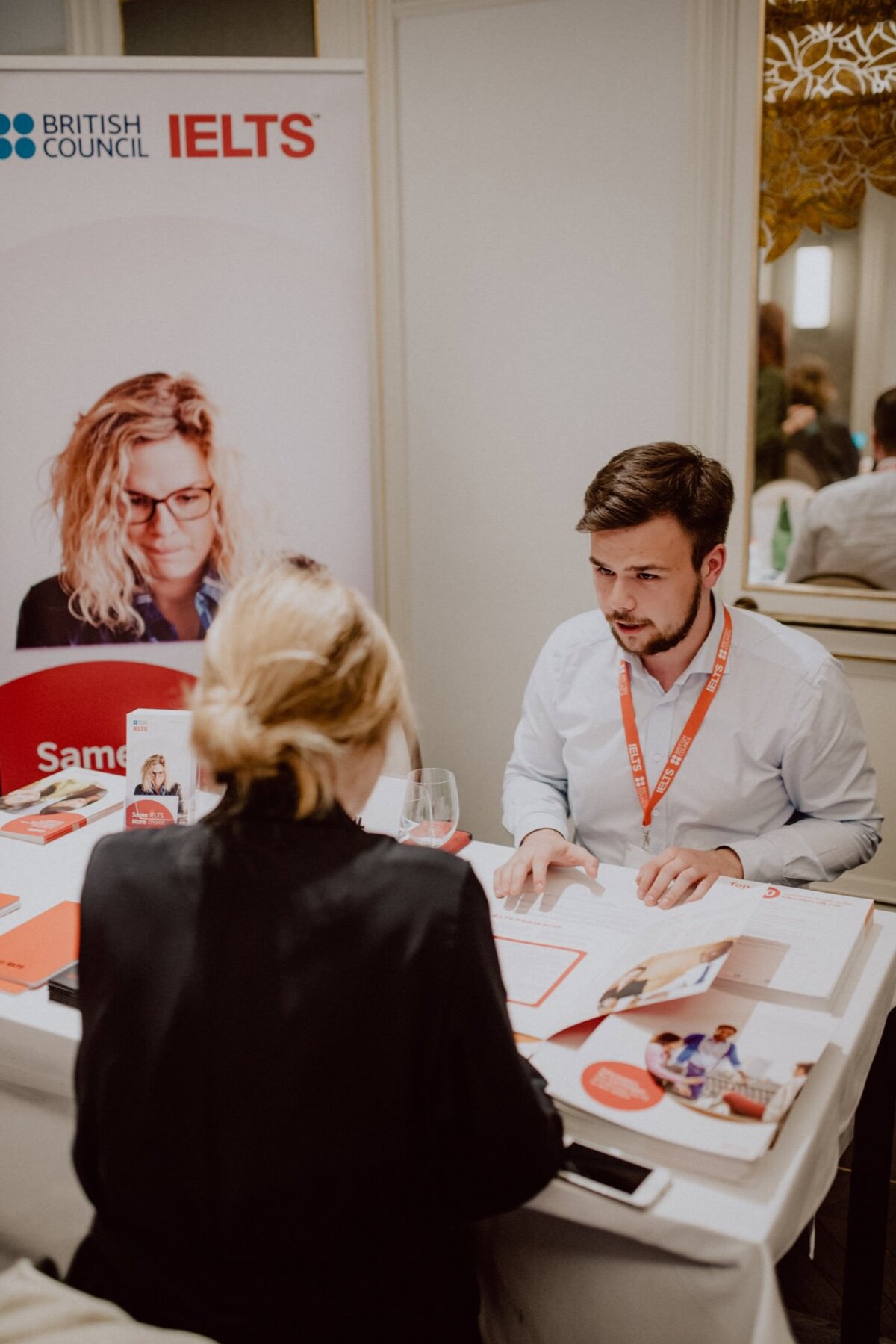 Two people are sitting at a table with IELTS and British Council logos. The person on the left is standing with his back to the camera, while the person on the right is busy talking and writing. The table is swamped with papers and promotional materials, perfectly capturing the photo coverage of the events.  