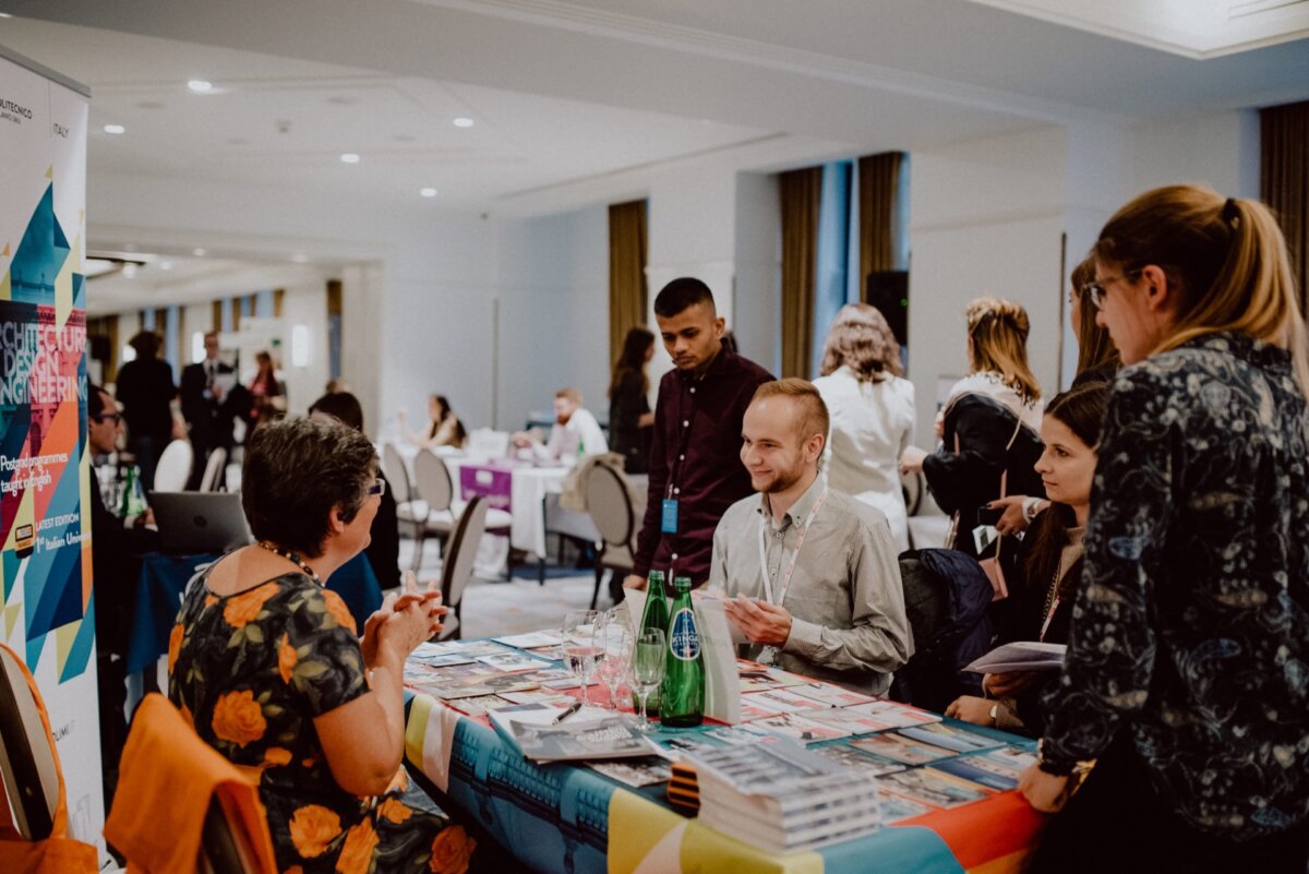 During an event held indoors, a group of people gather around a table. The table is covered with colorful brochures and a bottle of water. Participants, both seated and standing, are engaged in conversation. Other groups of people are also interacting in the background, capturing the perfect moment for event photography.   