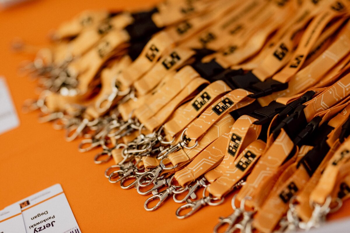 A stack of yellow lanyards with metal clips, marked with black and orange text, is spread on an orange surface. In the lower left corner you can see partially visible badges with text and logos resembling the lanyards - a perfect snapshot for event photography. 