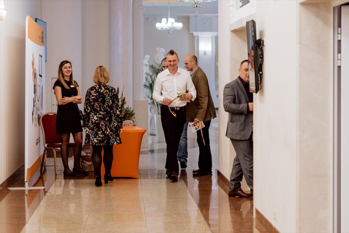 A well-lit hallway with beige walls and a polished floor. Five people are engaged in conversation at an orange-covered table of promotional materials. One man walks by in a white shirt holding a document, while another in a suit looks at a screen mounted on the wall, capturing the essence of event photography.  