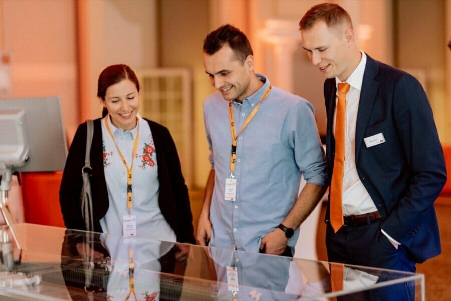 Three people with name badges and lanyards smile while viewing a display case at a brightly lit event. One man wears a suit; the others wear casual attire, captured by fotografia eventowa warszawa.