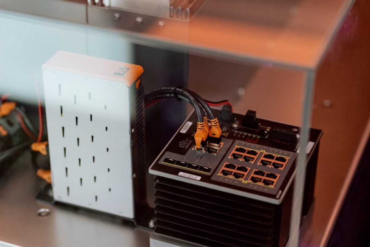 A close-up of a network switch and power supply enclosed in a glass case. The network switch has multiple Ethernet ports, some with cables attached. The equipment on display at the industrial conference is mounted on a metal surface, exposing technical and electronic components.  