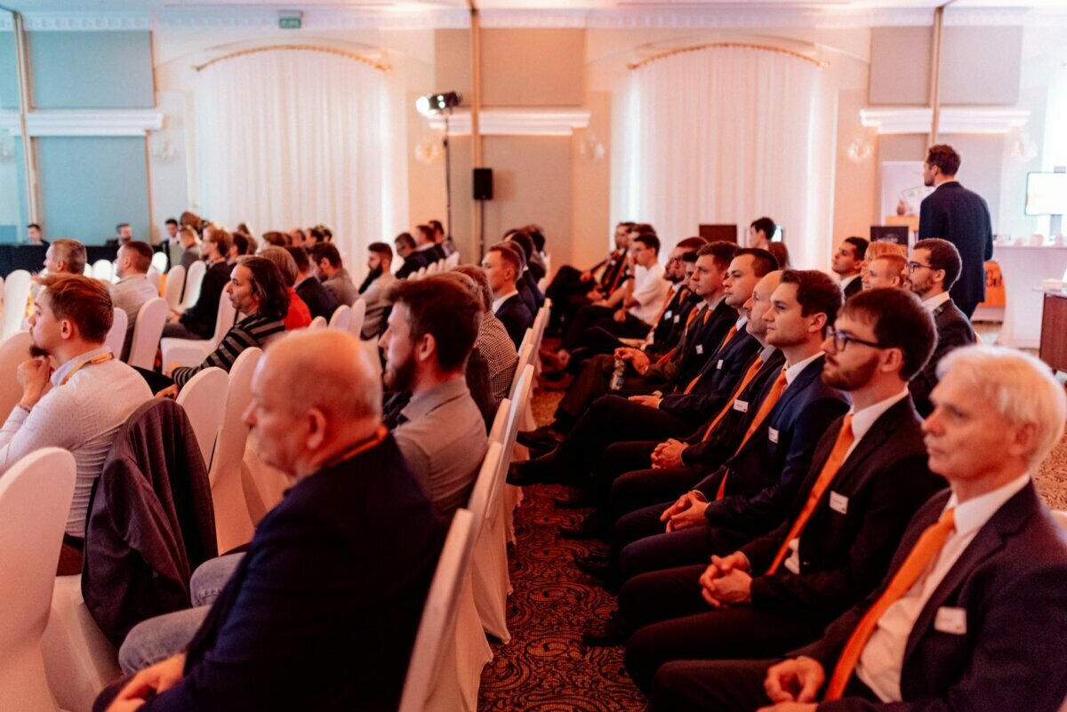 A group of people sitting in rows during a formal conference in a well-lit room. Many attendees are wearing suits and ties and listening intently to the presentation. The room is decorated with white curtains and ornate details, perfect for a professional event photographer Warsaw to capture photo coverage of the events.  