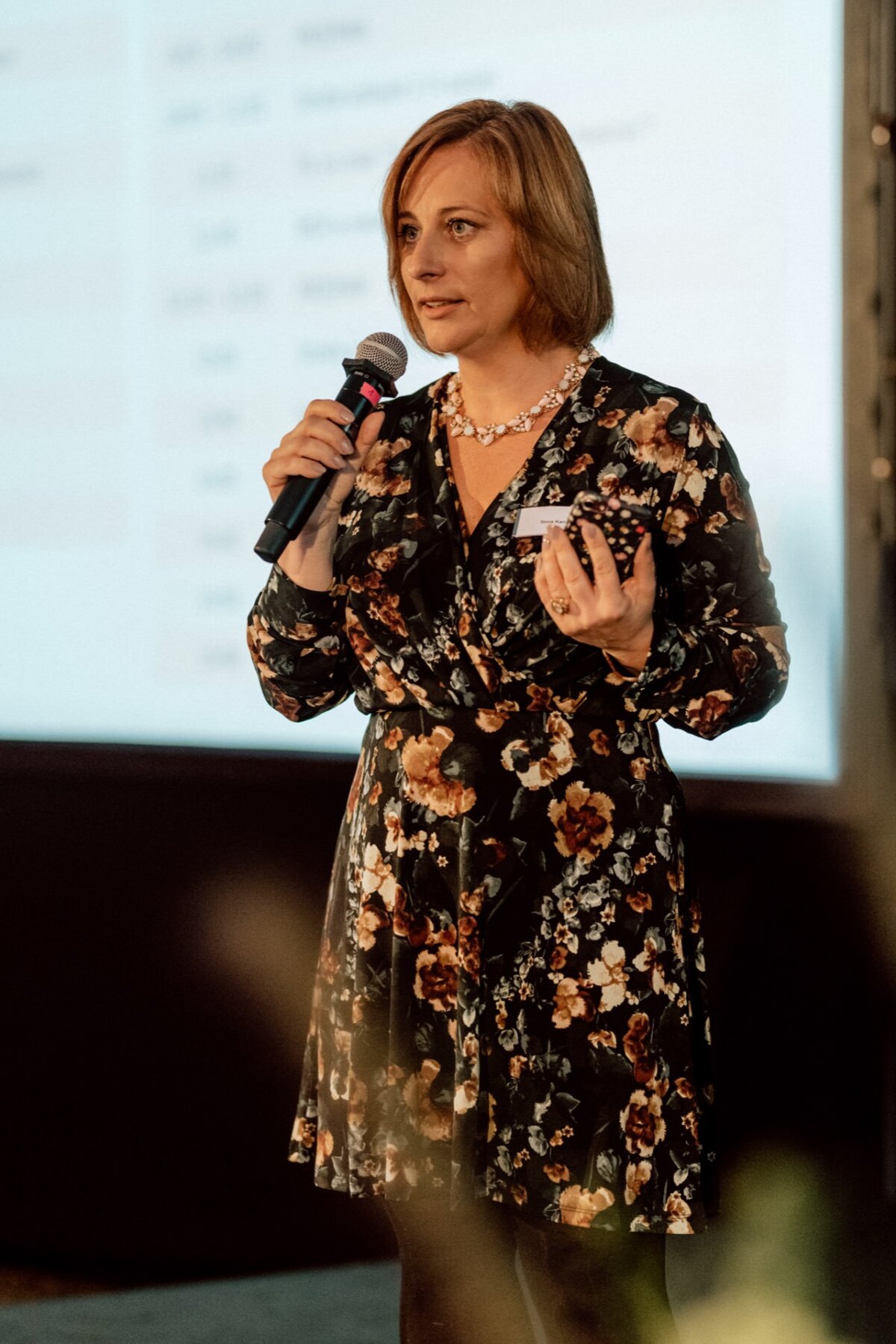 A woman in a flowery dress stands holding a microphone in her right hand and another object in her left. She speaks in front of a large screen or in the background of a presentation, apparently engaging the audience at the event. This scene captures the essence of event photography at its best.  