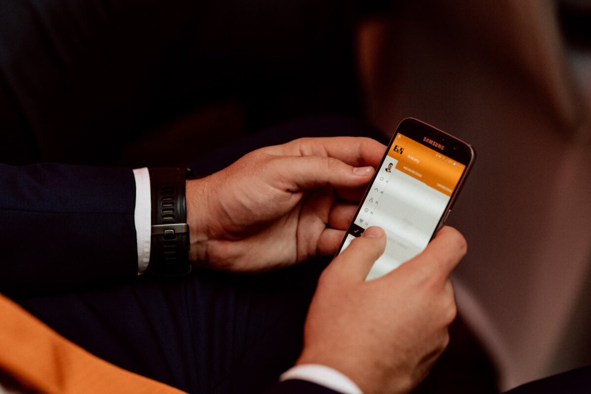 A person in a suit is holding a smartphone and navigating through a mobile application. The screen displays various options under a bright orange header, and the position of the person's hand suggests that he is about to touch a menu item. A watch can be seen on the person's left wrist - captured during a photo tour of the events.  