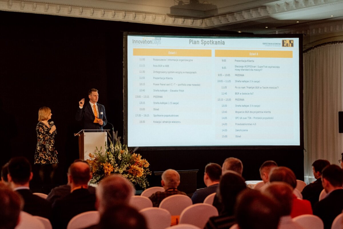 A man and a woman are speaking at a conference before a seated audience. Behind them, a large screen displays a detailed program titled "Meeting Plan." The room is well lit and the audience seems attentive, some using the equipment - an ideal location for event photography.  