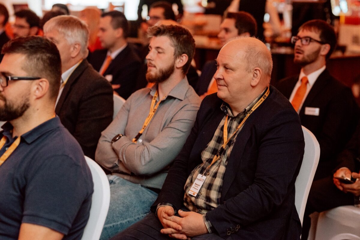 A group of men sitting in rows at a conference listen attentively to a presentation. They wear lanyards with badges and sports goggles. In the background is a blurry conference setting with people and a booth with various materials, captured by event photographer Warsaw.  