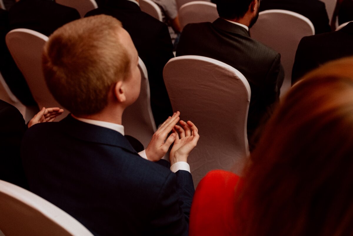 A person with short, light-colored hair, dressed in a dark suit, claps his hands while sitting among others at what appears to be a formal event. The people around him are similarly dressed in suits and are all seated in white-covered chairs - a captivating photo-illustration of the events that shows the elegance of the gathering. 