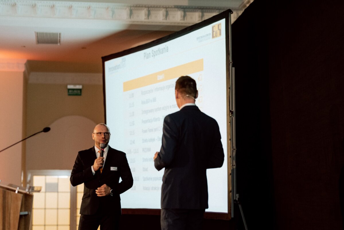 Two people in formal attire stand with microphones near a large screen displaying the presentation. The person on the left is facing the audience, the person on the right is with his back to the camera. In the background you can see a well-lit room and a section of the ceiling, perfect for event photography.  