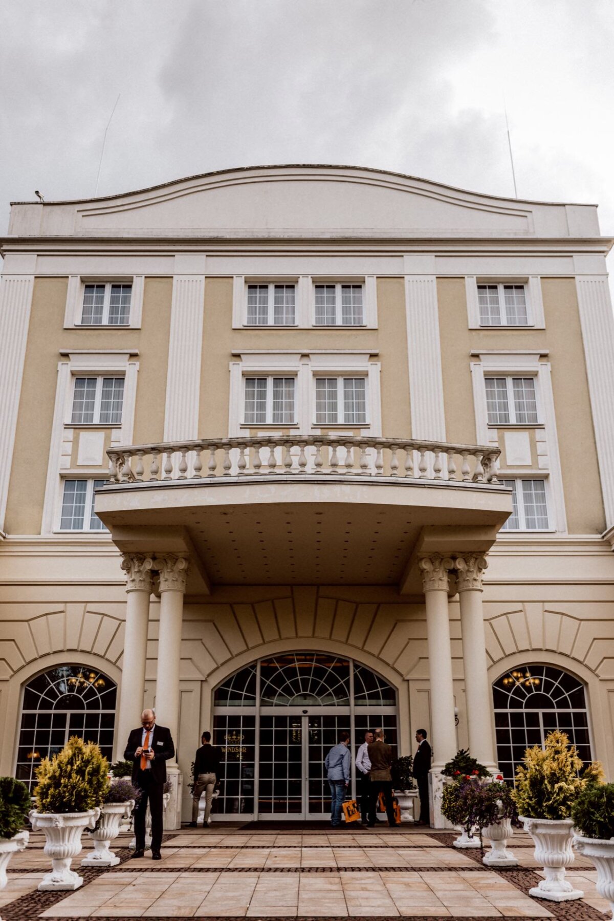 A stately building with large arched windows and columns at the entrance, adorned with a balcony above. Several people stand at the entrance, some chatting as if waiting for an event photo. Ornamental plants in white pots stand on either side of the path leading to the door. Above, an overcast sky.   