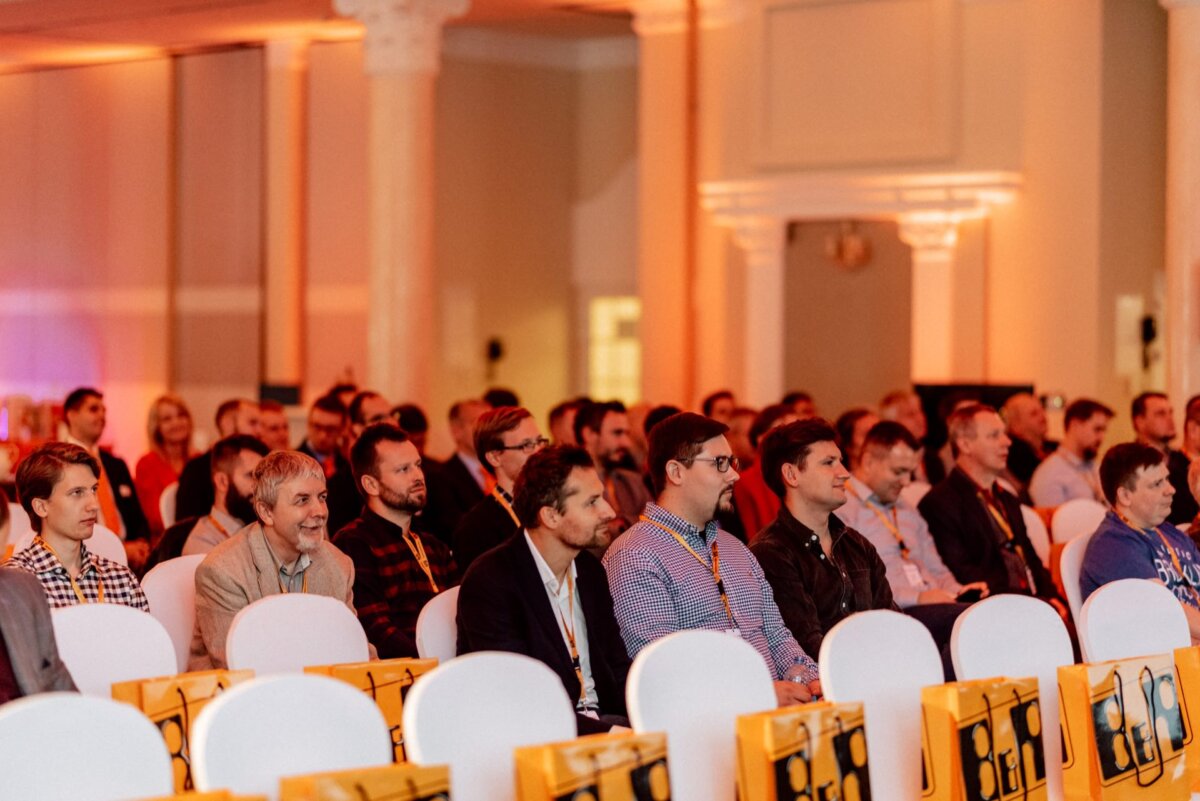 A large group of people sit in rows of white chairs and attend a conference or industry seminar in a warmly lit room. Many attendees are wearing conference lanyards around their necks, and yellow bags are stacked on some chairs. 