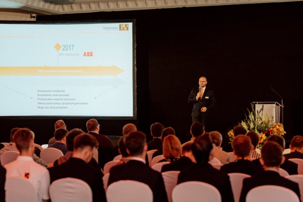 A man in a suit stands next to a podium and delivers a presentation to an audience seated in white chairs. A large screen displays a slide with text and graphics, including a timeline of 2017. Flowers arranged near the podium capture the essence of professional event photography.  