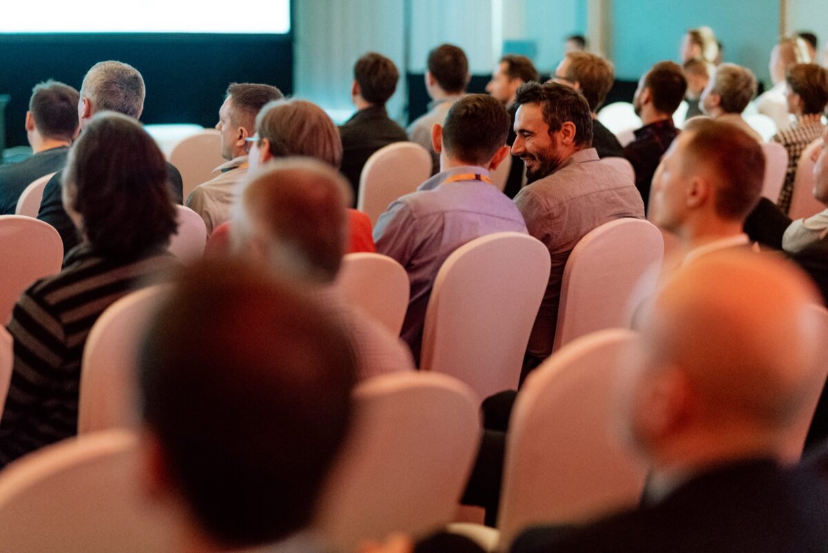 A group of people are sitting on white chairs in a conference room, facing a large screen at the front. Some of the participants are busy talking, while others are looking at the screen with interest. The atmosphere captured in the event photography gives the impression of being focused, yet relaxed.  