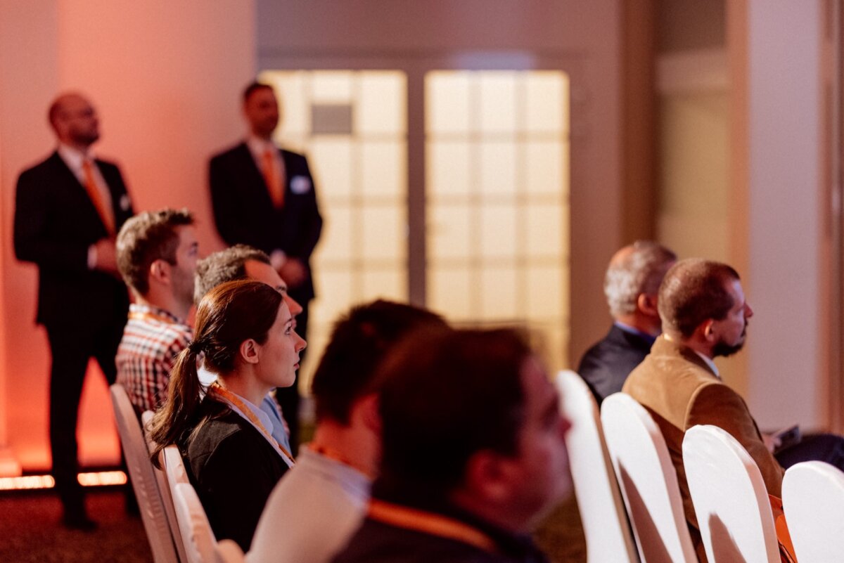 Several people are sitting in rows, carefully looking ahead in the well-lit conference room. Two people in suits stand at the back. Some attendees are wearing badges or lanyards. The atmosphere suggests a formal event or presentation, perfectly captured in this photo report of the event.   