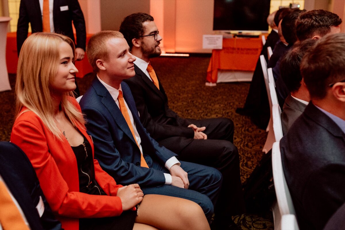 At an event held indoors, a group of people dressed in formal attire sit in rows. In the first row you can see two men and a woman, all dressed in orange outfits. They are looking intently ahead, with a screen and backlit display in the background - the perfect scene for event photography.  