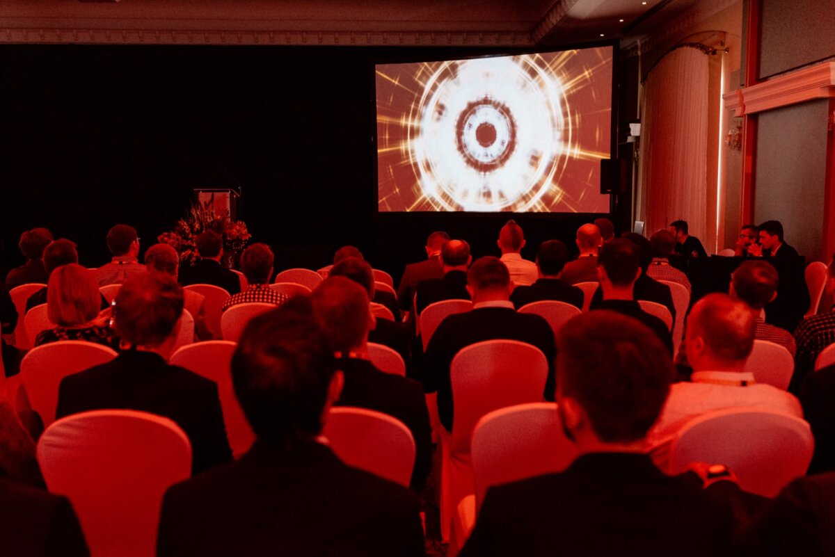 A group of people sitting in a darkened room, facing a large screen with a vivid circular display. The room is dimly lit with a red glow, and the audience gives the impression of attentively watching the presentation, perfectly captured by a specialist event photographer Warsaw. 