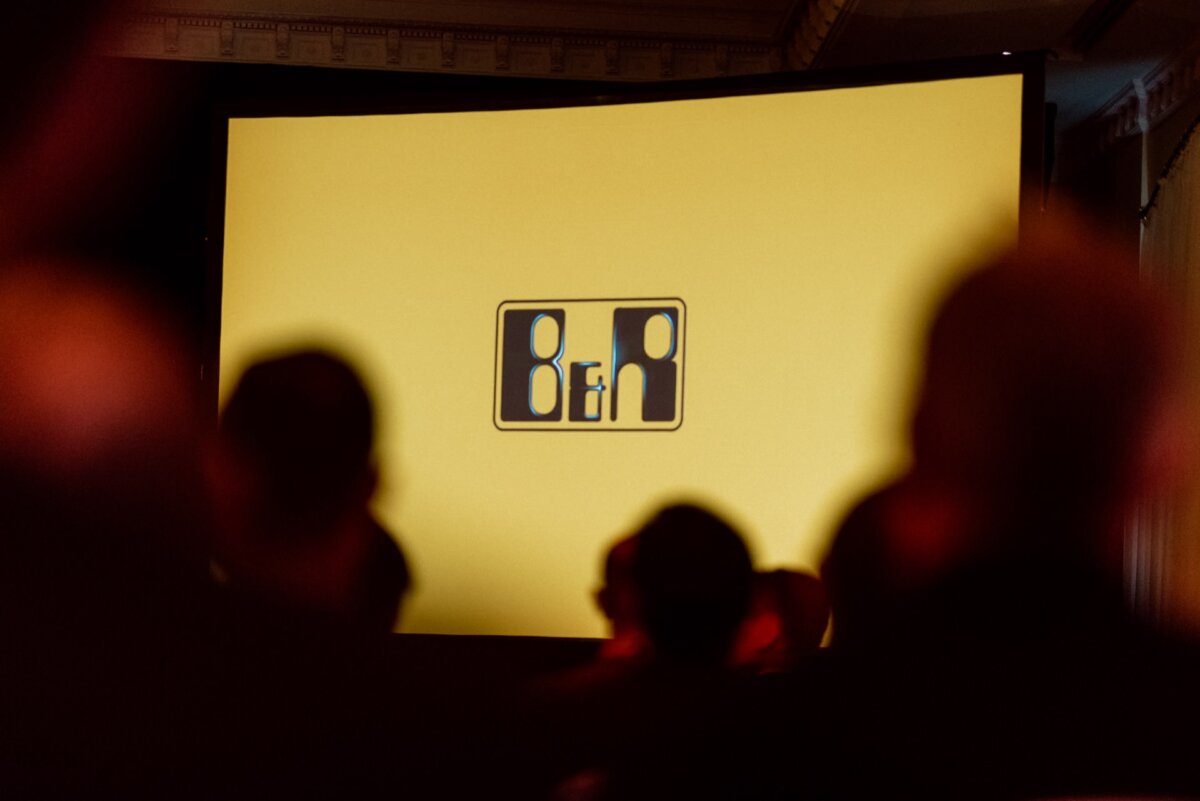 The projection screen displays a logo depicting three abstract human shapes: two larger ones on the sides and a smaller one in between. The background is yellow, and the silhouettes of the audience are visible in the foreground, capturing the essence of event photography. 