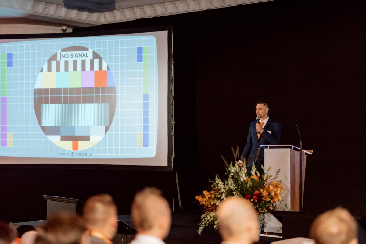 A person in a suit stands at a podium on the stage and gives a presentation. A large screen behind them displays a TV test pattern with the message "NO SIGNAL." The podium is decorated with a floral arrangement, and several audience members can be seen in the foreground - a perfect moment for photo coverage of the events.  