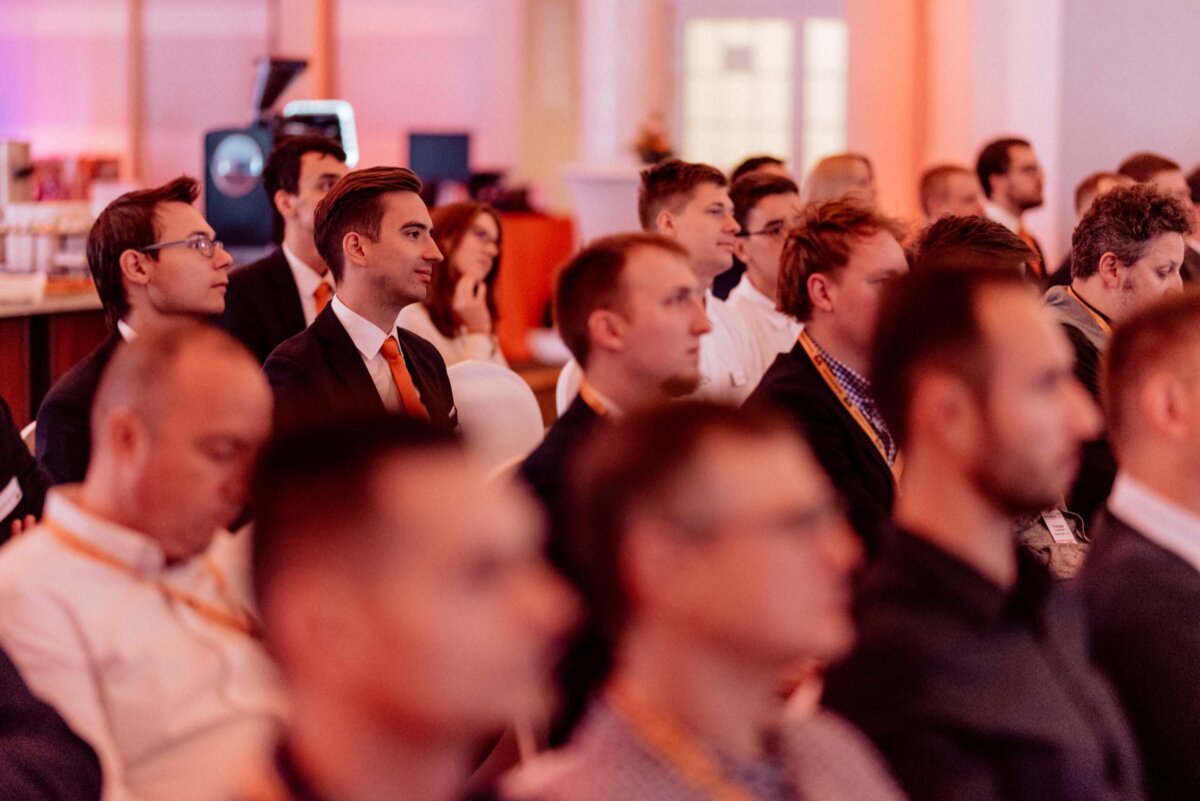 A group of people, mostly men, sitting attentively in rows at an event or conference held indoors. Some are wearing business attire with suits and ties, while others are wearing casual clothes. The atmosphere seems focused, with a projection screen visible in the background - perfect for an event photo shoot.  