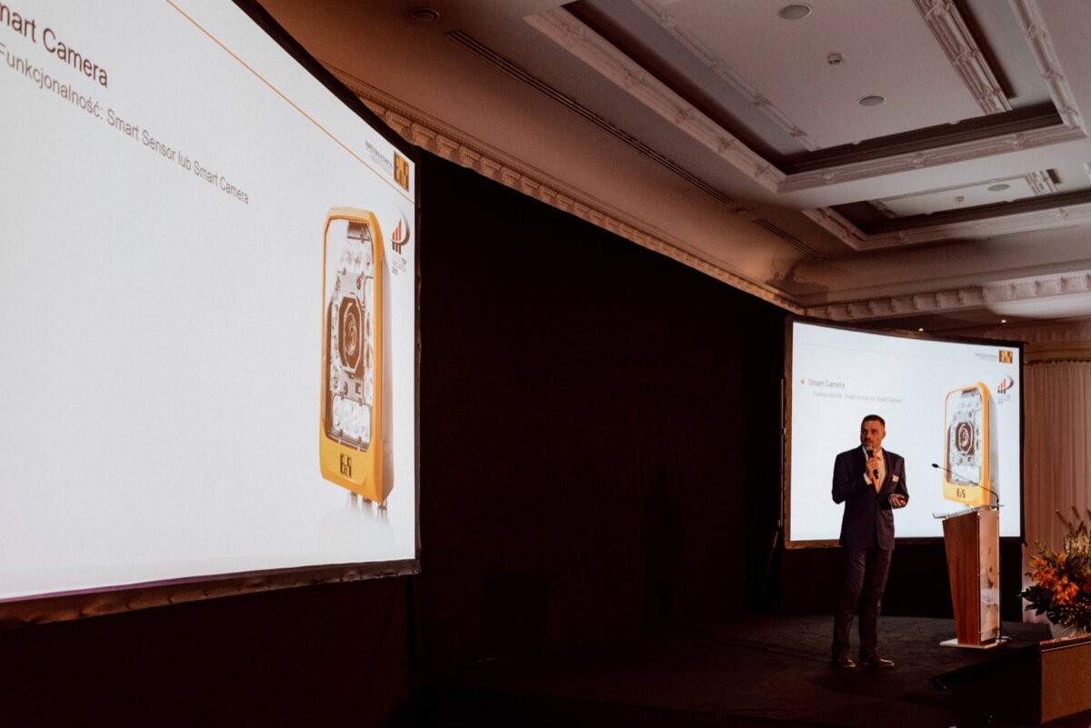 A person in formal attire stands on a stage and delivers a presentation in a conference room. The presentation slide behind them shows an image of a smart camera and text in German, perfect for event photography. To the right, another large screen can be seen displaying the same slide.  