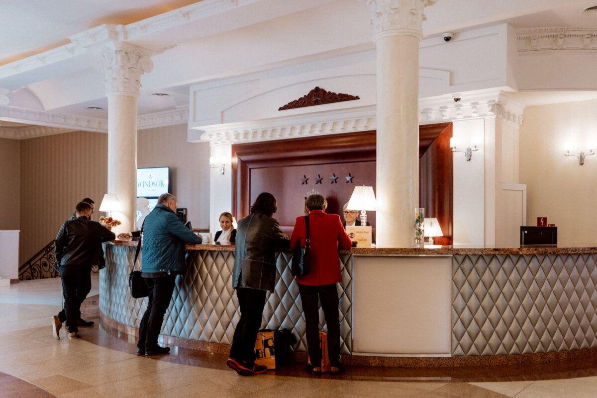 The hotel's reception desk with three people interacting with the staff behind the counter, captured in a beautiful event photo. The desk features elegant decor with quilted panels and marble surfaces. A staircase is visible to the left, and the overall atmosphere is luxurious and warmly lit.  