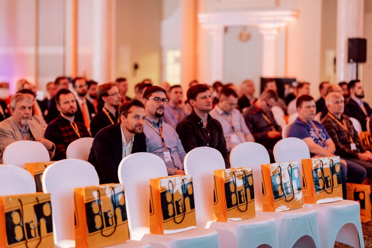 A large group of people sitting in rows of white chairs at an event or conference in a room. On some of the chairs in the foreground are orange and black gift bags. The atmosphere appears focused as the participants listen intently - the perfect moment for event photography.  