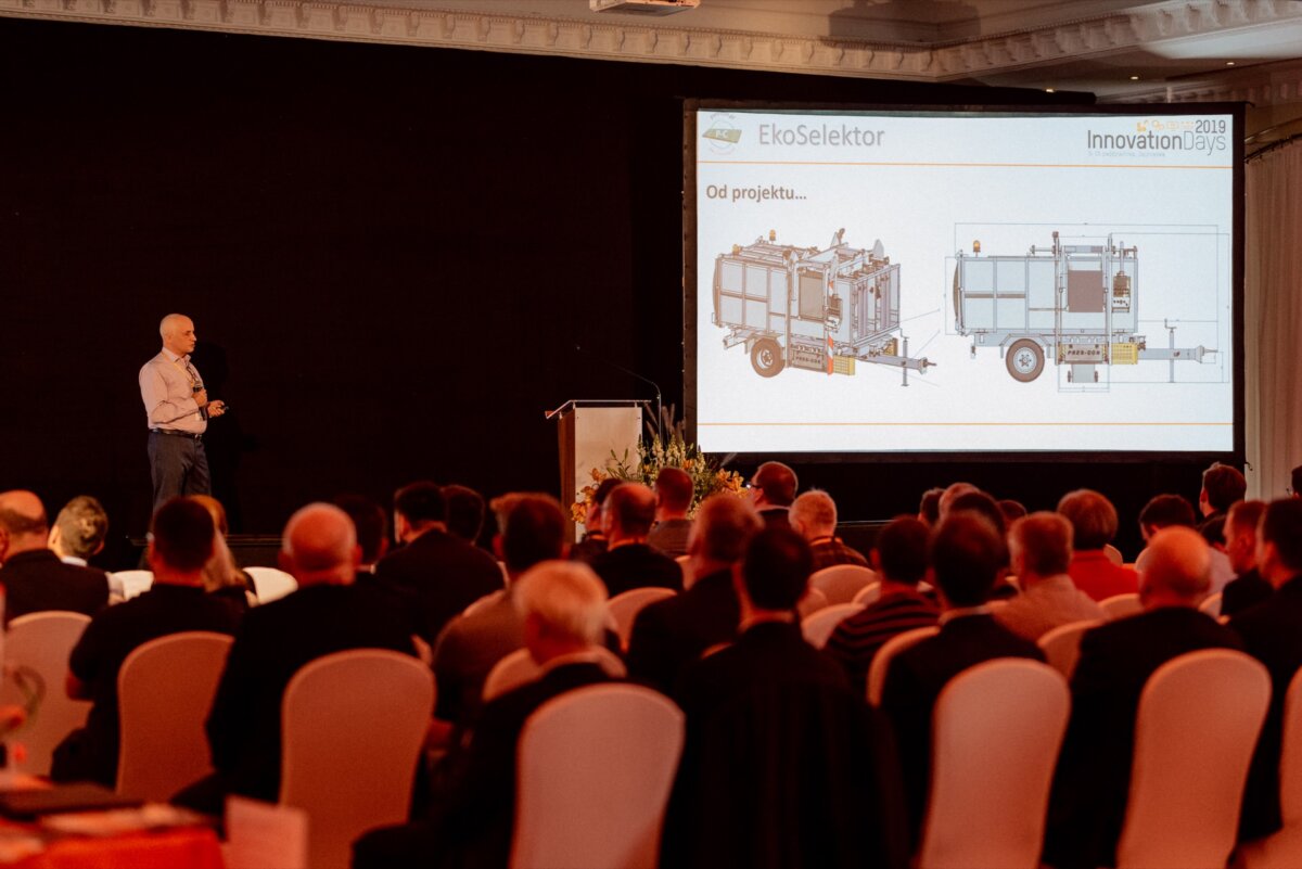 A man performs on stage in front of the audience, and behind him is a large screen displaying two diagrams of a machine with the words "EcoSelektor." The presentation is documented in a photo essay of the events taking place in the conference room. 