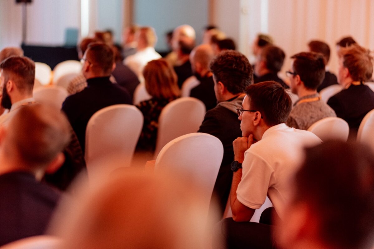 A group of people sitting in rows of white chairs and listening attentively to the speaker. Most of the people are facing forward, and attention is focused on the person in the white shirt in the foreground. This scene was beautifully captured as part of a photo report of events from what looks like a conference or seminar.  