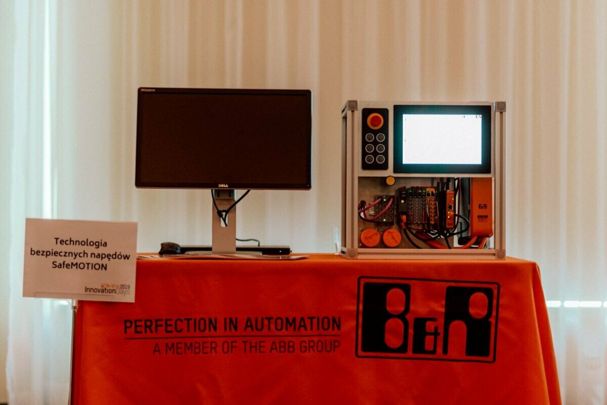 A display set consisting of a computer monitor next to an electric control panel mounted on a stand, placed on an orange tablecloth with the inscriptions "PERFECTION IN AUTOMATION" and "MEMBER OF THE ABB GROUP." The placard reads: "SafeMOTION safe drive technology." Ideal for event photography.  