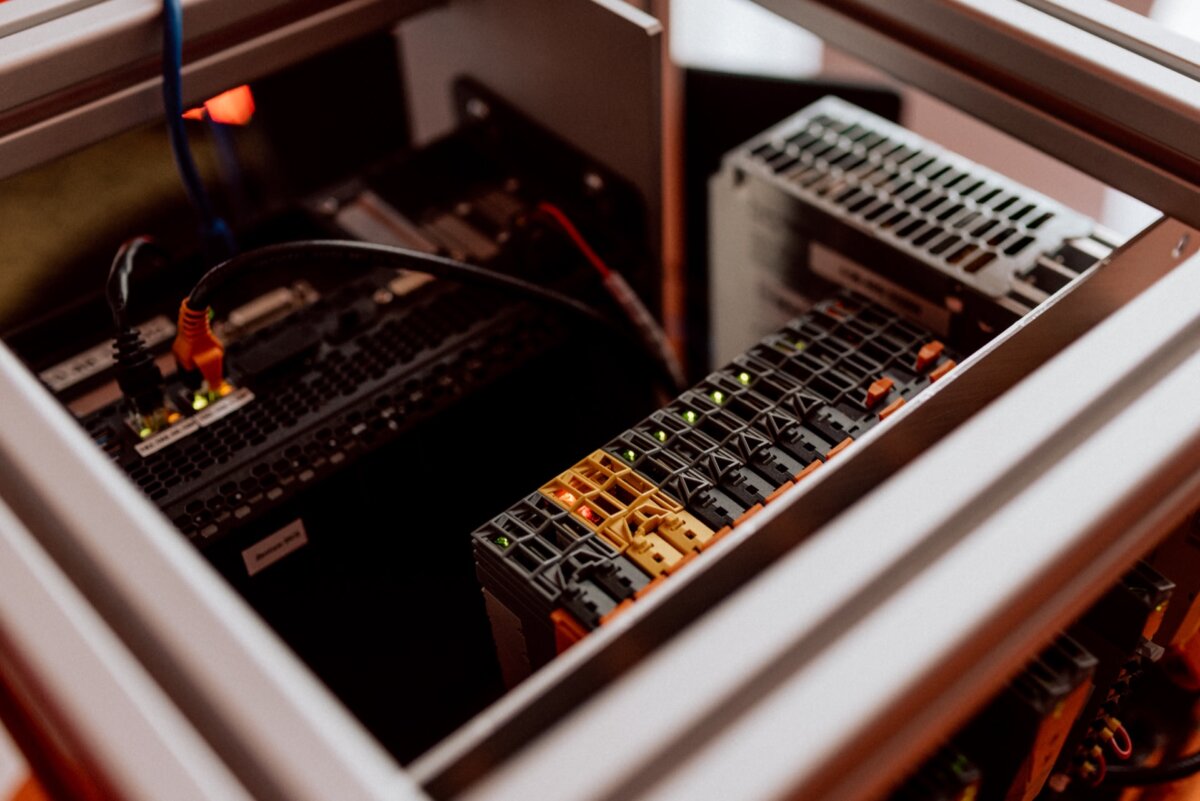 Close-up of a computer server rack containing various interconnected hardware components. Network cables and several server units with light indicators are visible, showing the data center infrastructure. The image, reminiscent of event photography, exposes electronic equipment in an orderly arrangement.  