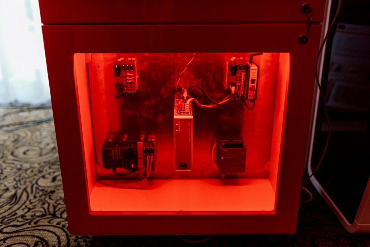 The open-front cabinet, illuminated by a red light, contains several electronic components and circuit boards mounted on the back panel. Wires connect the devices in the cabinet. The setup is reminiscent of the equipment you'd find when doing photo coverage of events, resting on a patterned carpet.  