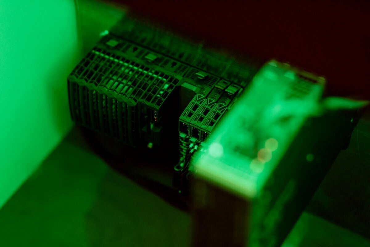 Close-up of server cabinets illuminated by green light, reminiscent of event photography. The photo depicts part of a server room or data center, showing multiple server units stacked and various wiring connections. The green hue gives the image a futuristic feel.  