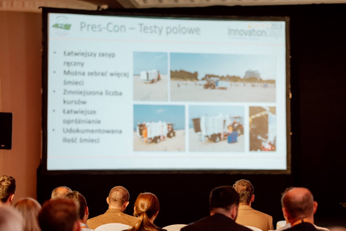 A group of people are seated in an auditorium, facing a large screen that displays a presentation slide in Polish with text and images. The slide appears to discuss the project with advantages and disadvantages, showing field tests and various configurations involving waste collection, all captured by event photography. 