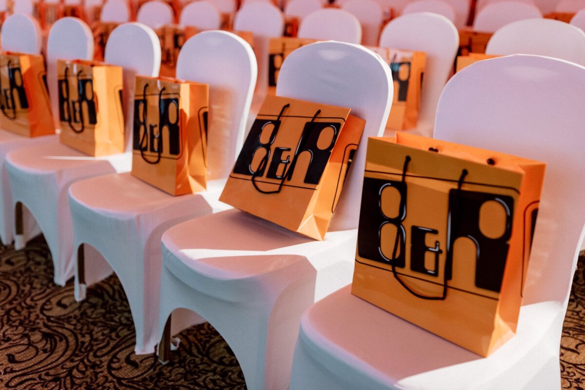 White chairs in a row, each with an orange gift bag with a black pattern placed on the seat. The chairs are arranged in a room lined with patterned carpeting, suggesting a conference or event setting. This event photo shows a well-organized arrangement awaiting attendees.  