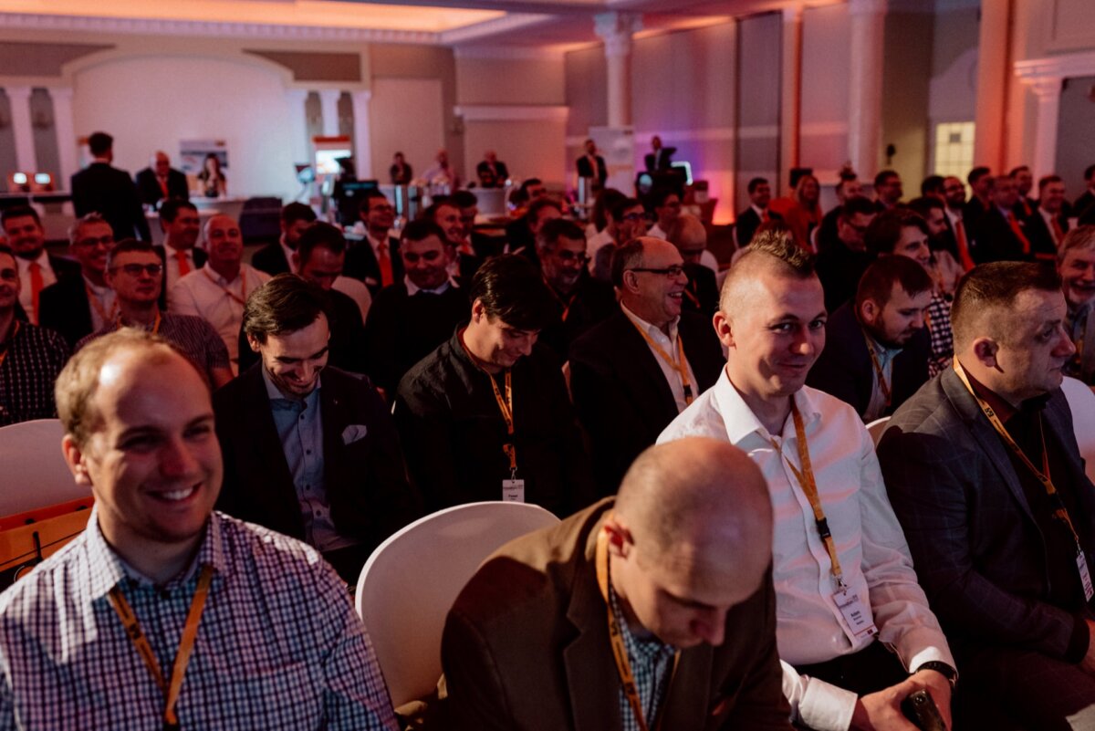 A large group of men in business attire sit in rows attending a conference or seminar in a spacious, well-lit room. Some are smiling and seem engaged, while others are looking down or to the side. A lanyard is visible on a few of the attendees, as captured perfectly by the event photography.  
