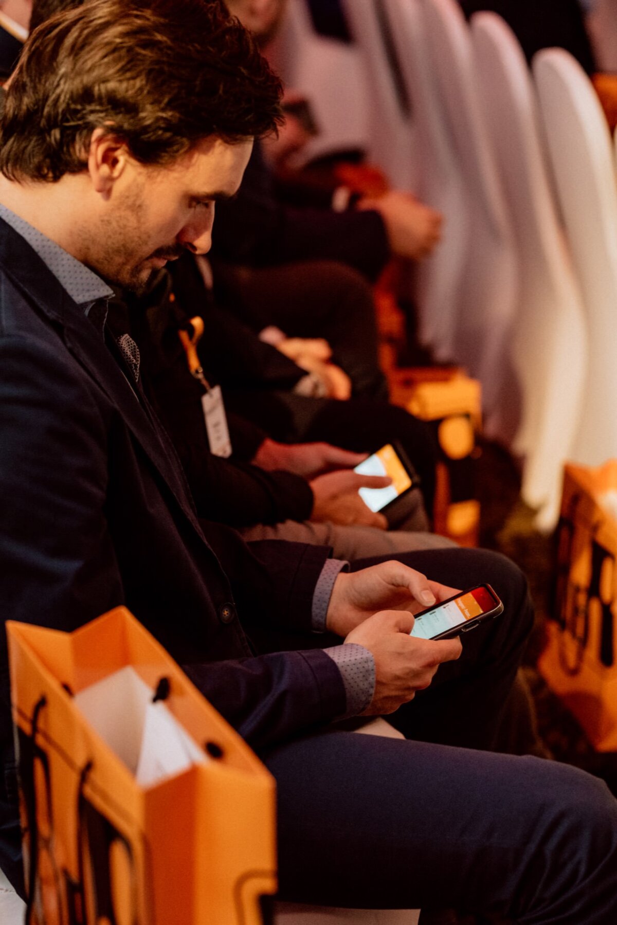 In a row of chairs sits a man with brown hair and a beard, looking at his smartphone. He is wearing a dark jacket and a light-colored shirt. Other people nearby are also using their phones. Brightly colored bags are placed next to the seats, illustrating a vivid photo essay of the ongoing events.   