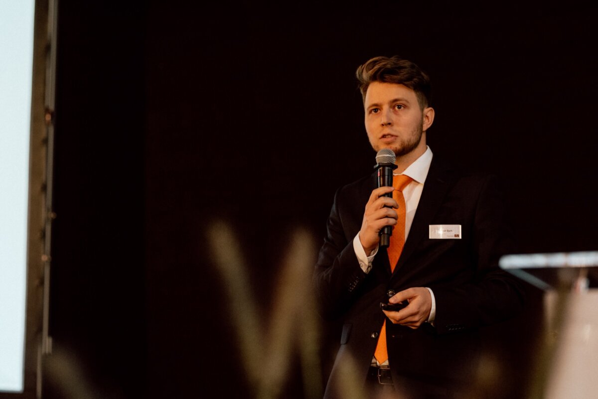 A man in a black suit, white shirt and orange tie stands with a microphone and small device and speaks during an event. The photo taken by event photographer Warsaw has a dark background with a backlit screen, partially visible on the left. 