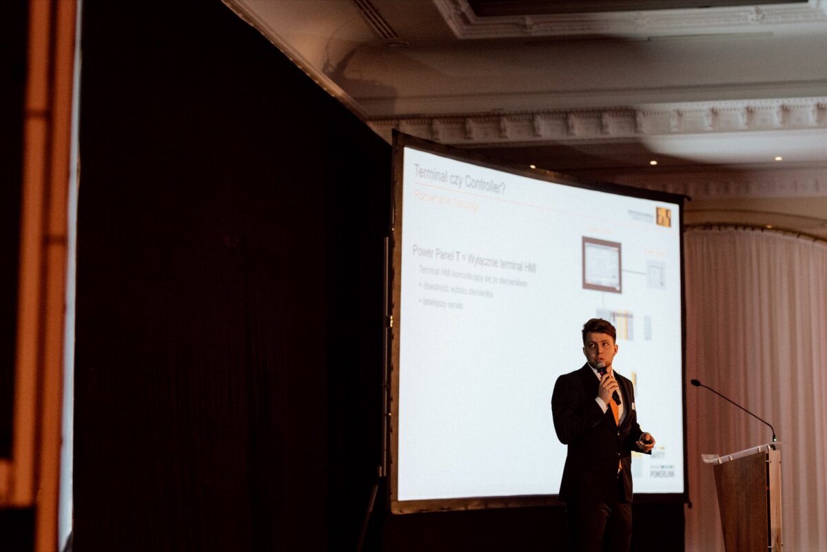 A person in a suit stands on stage, holds a microphone and performs in front of a backlit screen. The backdrop is a curtain and an ornate ceiling. The screen features technical-themed text and diagrams, perfect for an engaging photo essay of the event.  