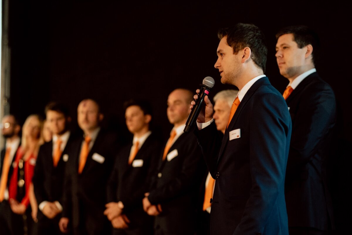 A man in a suit speaks to the audience, holding a microphone, and behind him against a dark background is a row of formally dressed men and women in black suits with orange ties. This scene could be part of an elegant event photo essay captured by an event photographer warszawa. 