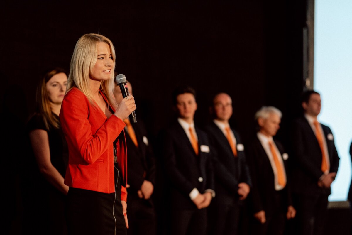 A blonde woman in a red jacket speaks into a microphone on stage. Behind her, several men and women in business attire are lined up, all facing forward. The background is dark, with an illuminated screen to the right - beautifully captured by an event photographer Warsaw for event photojournalism.  