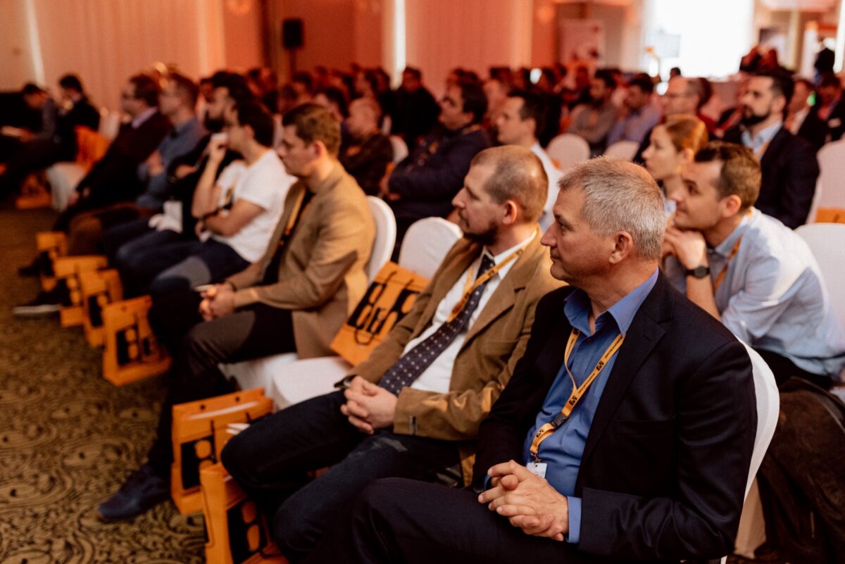 Spectators, mostly men in business attire, sit attentively in the conference room. Some have yellow lanyards and orange bags. The room is well-lit and filled, with a patterned carpet and white chairs. The background shows more attendees and a bright entrance - it really is the perfect scene for event photography.   