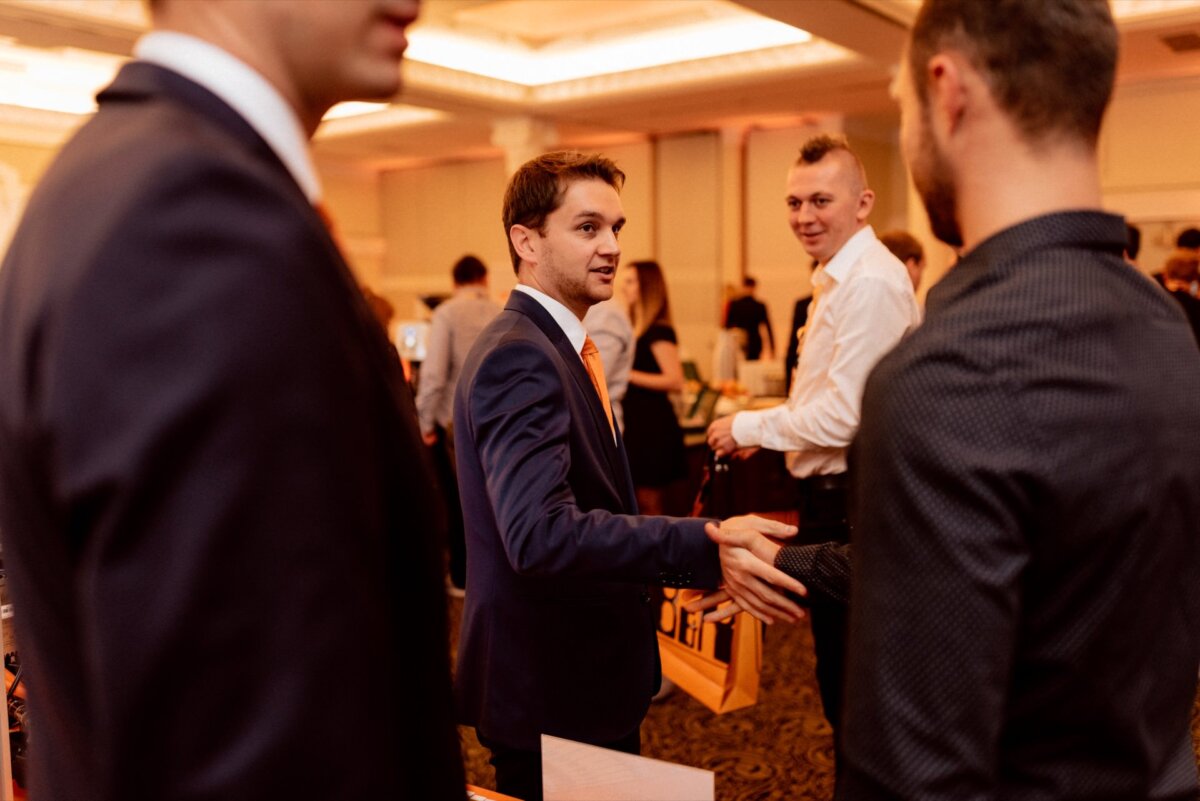 A man in a blue suit and orange tie is shaking hands with another person in a well-lit room. Nearby are other people dressed in business or formal attire, engrossed in conversation. The backdrop appears to be a professional or networking event, ideal for anyone who wants to capture a photo-essay of events.  