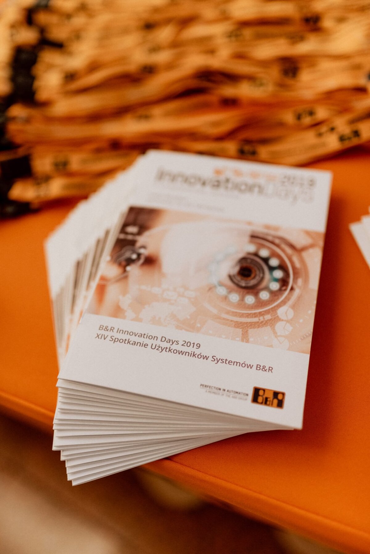 On an orange table is neatly stacked a stack of brochures titled "B&amp;R Innovation Days 2019 XIV B&amp;R Systems User Meeting." The cover design shows a close-up graphic of machines and gears. Yellow lanyards can be seen in the background, perfectly capturing the essence of event photographer warszawa.  