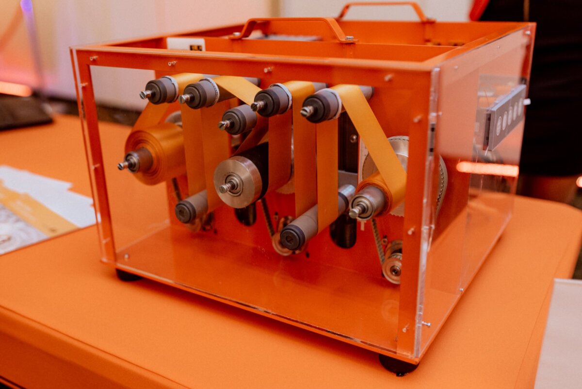 A close-up of a machine in a transparent orange enclosure containing multiple spools and reels, captured in a photo report of the events. The machine appears to be designed for processing or transferring materials such as ribbons or tape. It is placed on an orange table.  