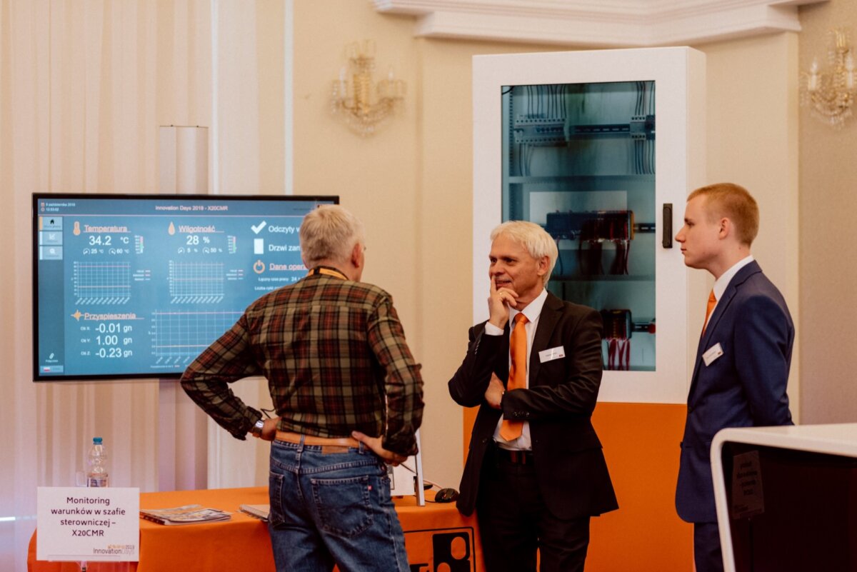 Three men are discussing at a technology booth. On the left, a man in a checkered shirt faces two men in suits. A large screen displays technical charts and the data behind them, clearly captured by an event photographer Warsaw at this exhibition or conference.  